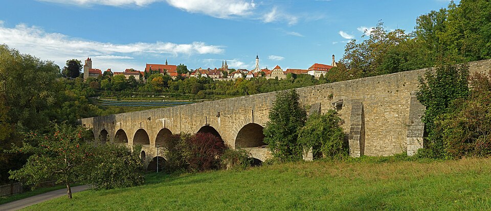 The walled town of Rothenburg ob der Tauber seen across the green Tauber valley with a double-arched bridge below.